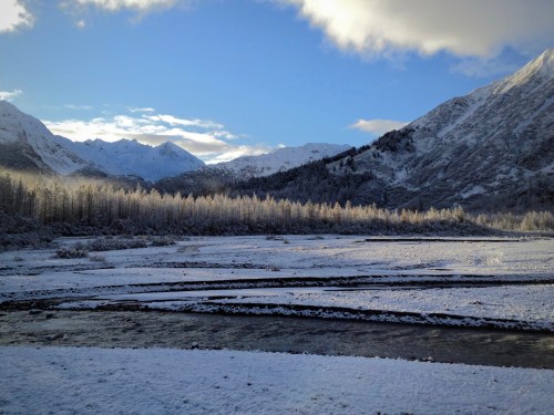A snowy landscape with mountains in the background, a river flowing through, and trees covered in frost under a blue sky.