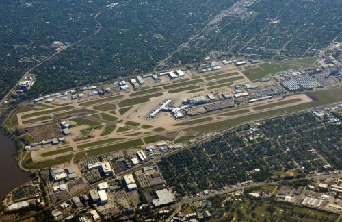 Aerial view of a large airport with runways, taxiways, and surrounding green areas and buildings.