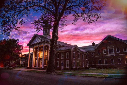 A historic building with columns, surrounded by trees, under a vibrant sunset sky with colorful clouds.