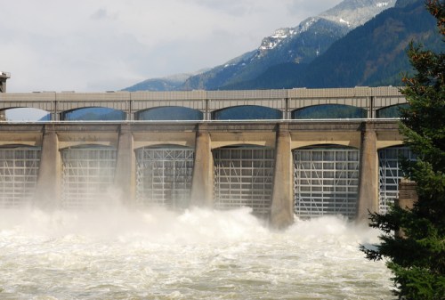A large dam with multiple gates releasing water, surrounded by mountains and trees under a cloudy sky.