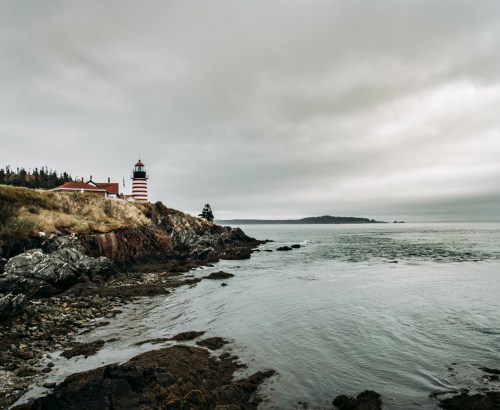 A lighthouse stands on a rocky shore under a cloudy sky, overlooking a calm sea and distant land.