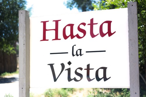 Sign reading "Hasta la Vista" in bold red and black letters against a white background, surrounded by greenery.