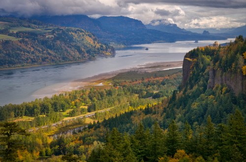 A scenic view of a river winding through lush green hills and forests, with mountains in the background under a cloudy sky.