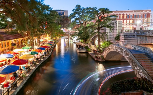 A scenic view of a riverwalk at dusk, featuring colorful umbrellas, boats, and illuminated buildings along the water.