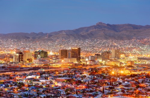 A panoramic view of a city at dusk, with illuminated buildings and mountains in the background.