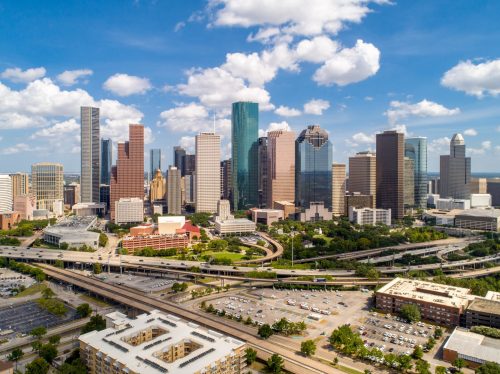 Aerial view of Houston's skyline with tall buildings, highways, and scattered clouds in a blue sky.