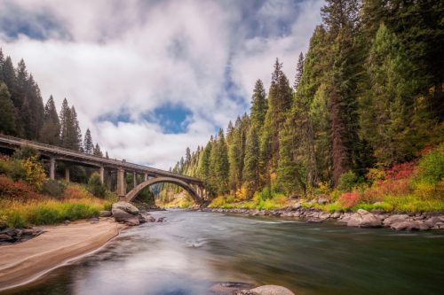 A serene river flows under a bridge, surrounded by lush green trees and colorful autumn foliage against a cloudy sky.