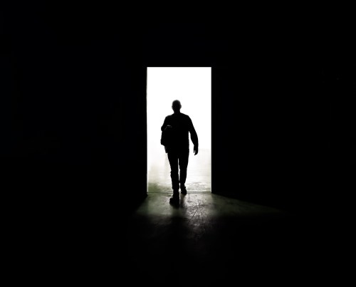 A silhouette of a person walking through a brightly lit doorway against a dark background.