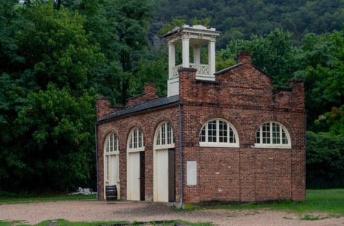 A brick building with arched windows and a bell tower, surrounded by lush greenery and gravel ground.