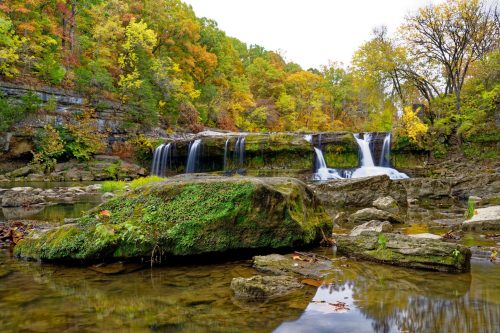A serene waterfall cascades over rocks, surrounded by vibrant autumn foliage and a calm reflective pool.