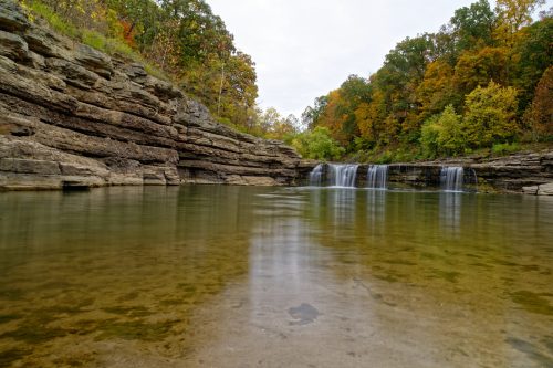 A serene landscape featuring a calm pool of water with a waterfall surrounded by colorful autumn foliage.