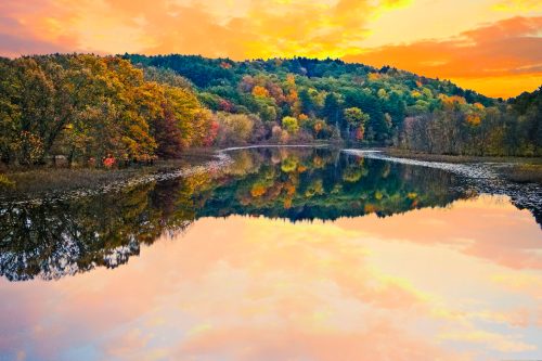 A serene lake reflects vibrant autumn trees and a colorful sunset sky, creating a peaceful natural scene.