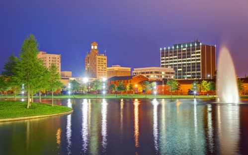 City skyline at dusk, featuring a clock tower, modern buildings, and a fountain reflecting in a calm lake.