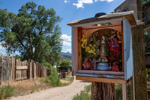 A small shrine with flowers inside, set along a dirt path surrounded by trees and a wooden fence.