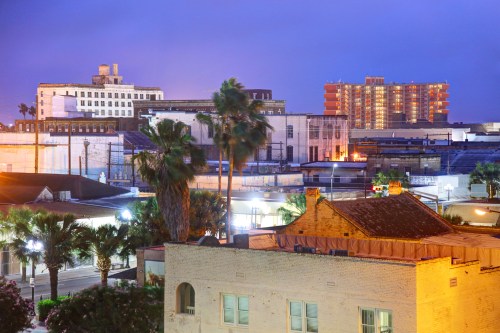 Cityscape at dusk featuring palm trees, historic buildings, and modern architecture under a purple sky.