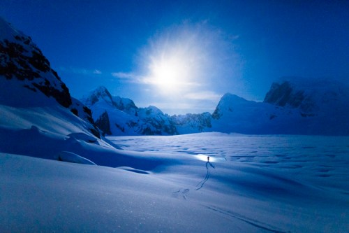 A lone skier traverses a snowy landscape under a bright moon, surrounded by majestic mountains.