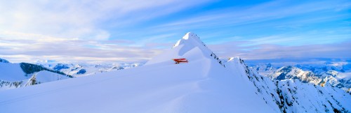 A red cabin sits atop a snowy mountain peak, surrounded by vast, snow-covered landscapes and a blue sky.