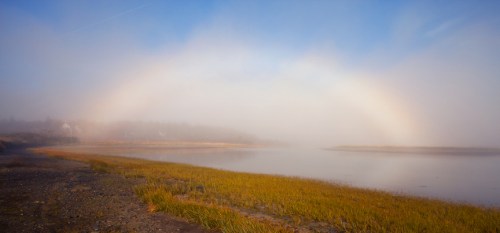 A soft rainbow arches over a misty landscape, with a calm river and grassy shore under a blue sky.