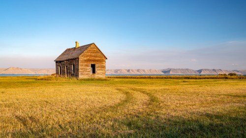 A weathered wooden house stands alone in a grassy field, with mountains and a lake in the background under a clear sky.