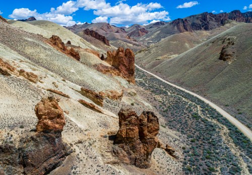 Aerial view of rugged mountains and a winding dirt road through a dry, rocky landscape under a blue sky with clouds.