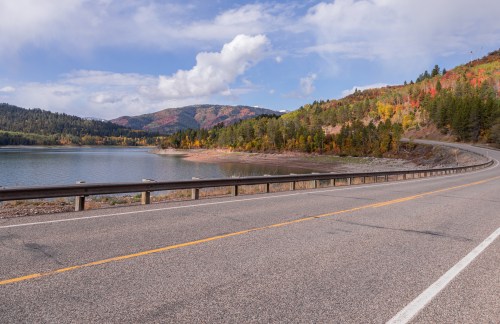 A winding road alongside a calm lake, surrounded by colorful autumn foliage and mountains under a partly cloudy sky.