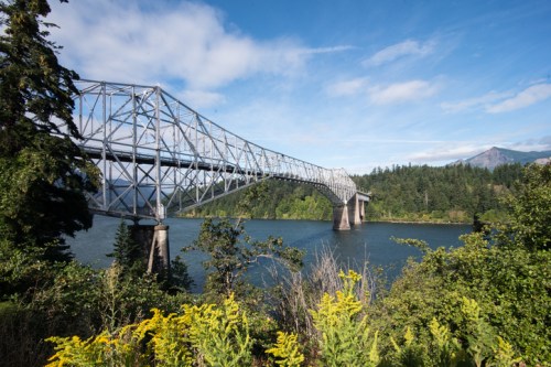 A steel bridge spans a river, surrounded by lush greenery and mountains under a blue sky with scattered clouds.