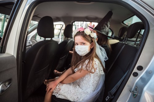 A young girl in a white dress and mask sits in the backseat of a car, wearing a floral headband and looking at the camera.