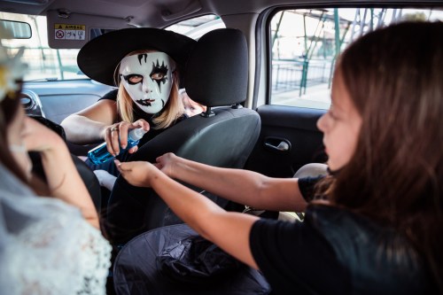 A person in a witch costume with a mask hands a spray bottle to a child in a car.