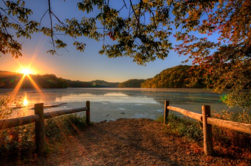 Sunrise over a tranquil lake, framed by trees and a wooden fence, with soft light reflecting on the water's surface.