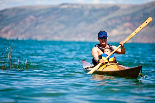 A person and a child paddle a kayak on a calm lake surrounded by mountains and greenery.