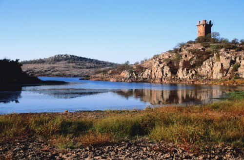 A serene landscape featuring a tower on a rocky hill beside a calm lake, surrounded by greenery and clear blue skies.