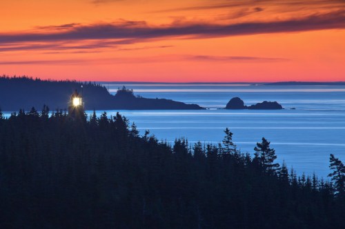 A lighthouse shines at sunset, surrounded by dark trees and calm waters, with vibrant orange and blue skies.