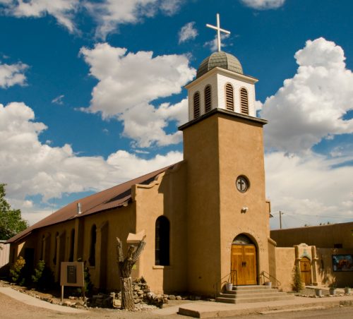 A rustic church with a cross atop its tower, set against a blue sky with fluffy clouds.