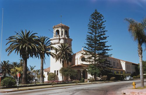 A sunny street view featuring a historic building with a bell tower, surrounded by palm trees and lush greenery.