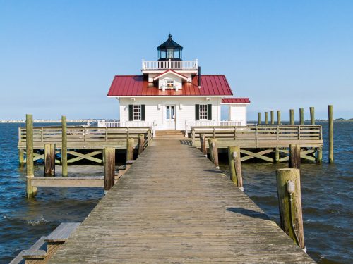 A wooden pier leads to a white lighthouse with a red roof, surrounded by calm water and a clear blue sky.
