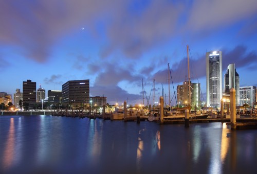 City skyline at dusk with boats docked in a marina, reflecting lights on calm water under a cloudy sky.