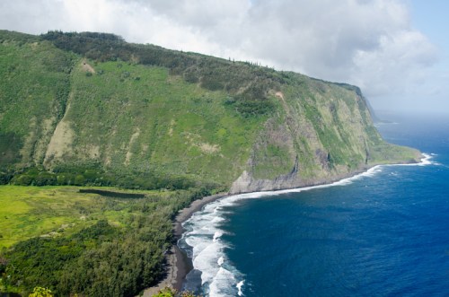 Lush green cliffs meet the ocean, with waves crashing on a black sand beach under a partly cloudy sky.