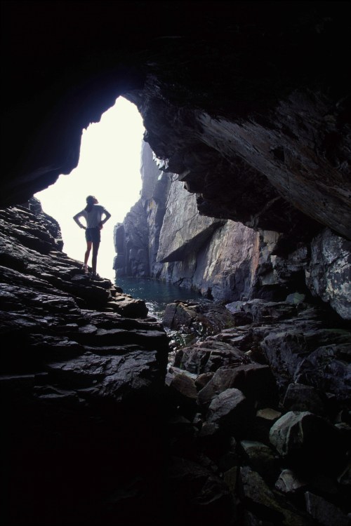 A silhouette of a person standing in a rocky cave, overlooking a serene body of water and towering cliffs.