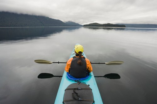 A person in a kayak paddles on calm water, surrounded by mountains and overcast skies.