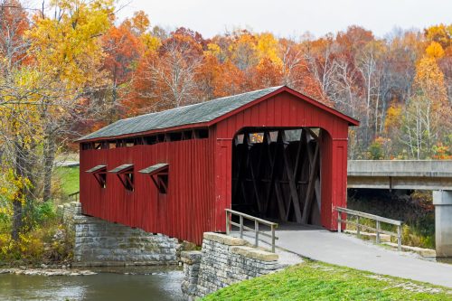 A red covered bridge over a stream, surrounded by vibrant autumn foliage in shades of orange and yellow.