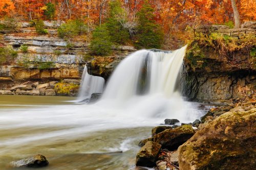 A serene waterfall cascades over rocks, surrounded by vibrant autumn foliage in warm hues of orange and yellow.