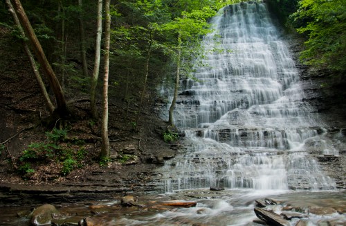 A serene waterfall cascading down rocky steps, surrounded by lush green trees and a tranquil stream below.