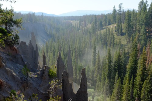 A misty valley with towering rock formations surrounded by dense evergreen trees under a clear blue sky.