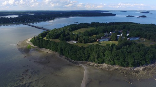 Aerial view of a lush green island surrounded by calm waters and a distant bridge, with scattered clouds in the sky.