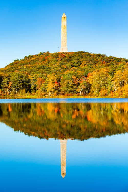 A tall monument on a hill reflects in a calm lake, surrounded by colorful autumn trees under a clear blue sky.