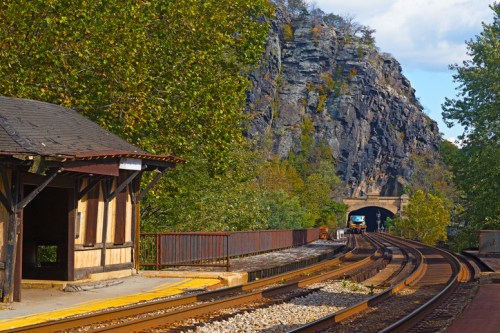 A train station beside a rocky cliff, with tracks leading into a tunnel under a blue sky and scattered clouds.