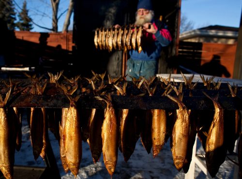 A person with a beard stands behind a rack of smoked fish hanging outdoors in winter.