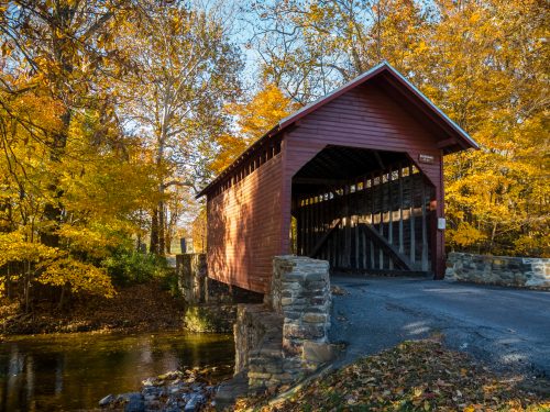 A red covered bridge surrounded by vibrant autumn foliage and a calm stream.