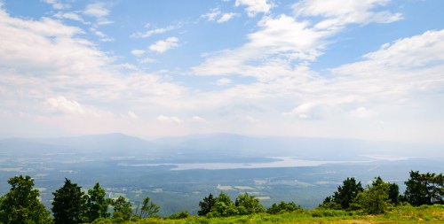 A panoramic view of a valley with a lake, surrounded by green hills and a blue sky with scattered clouds.