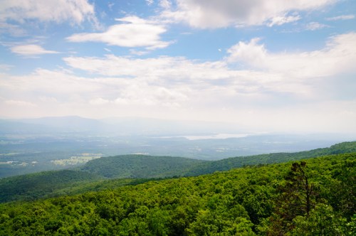 A panoramic view of lush green mountains under a partly cloudy sky, with a distant lake visible in the background.